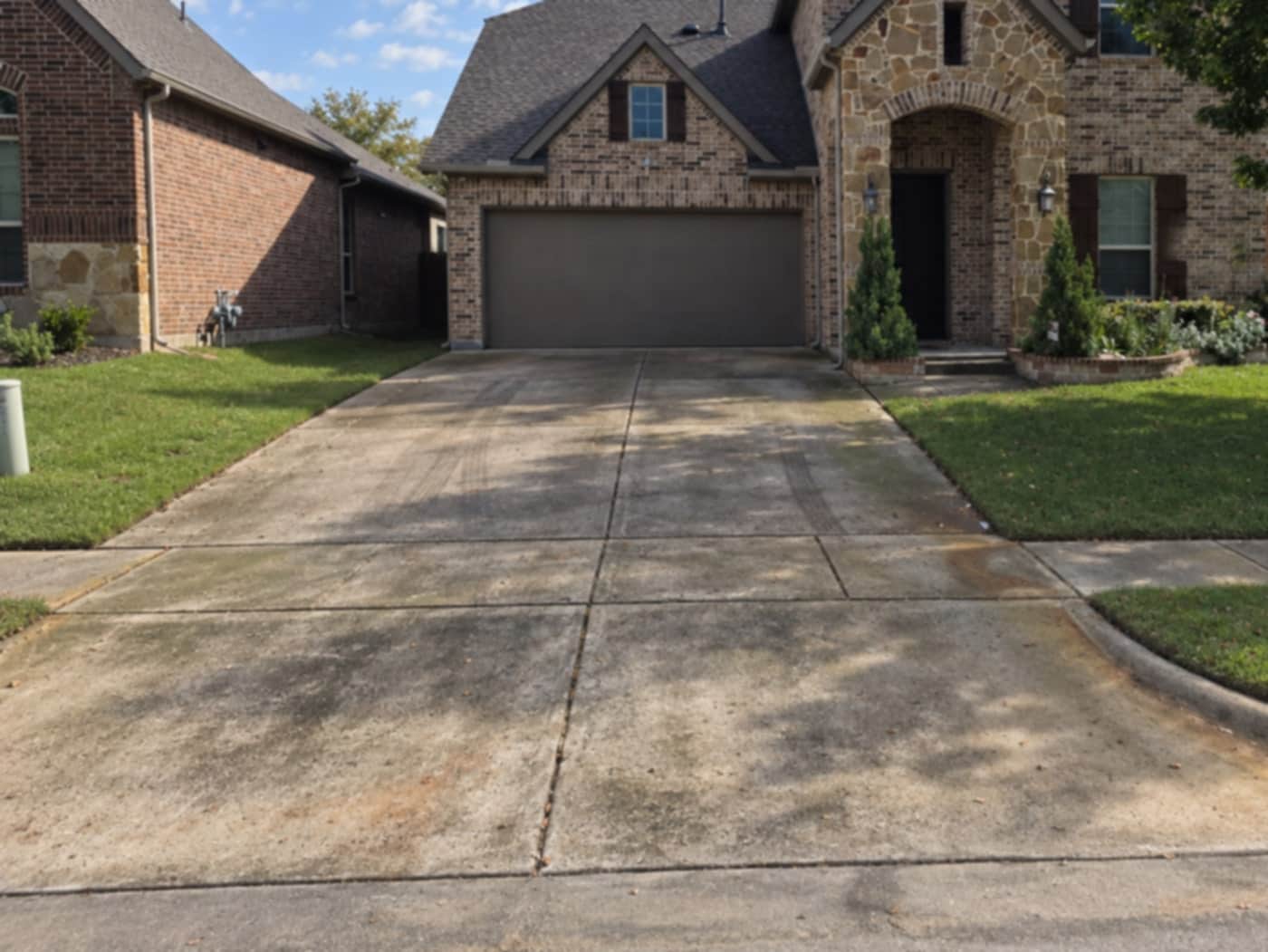 Concrete driveway of a two-story brick and stone home, grey and streaked with tire tracks and organic staining before cleaning.